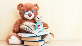 A large teddy bear in glasses holding a small teddy bear and reading from a pile of books.