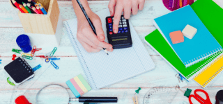 Table scattered with notebooks, erasers, pencils, paperclips, and other study supplies. In the center, a pair of hands is using a calculator and graphing paper.
