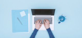 Hands typing on a laptop against a blue background. A mug of blue pencils and a blue notebook are arranged beside the laptop.